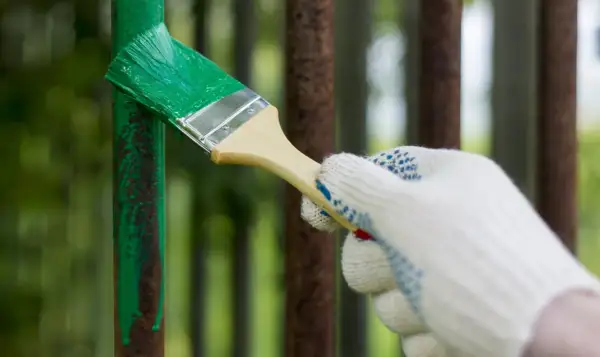 Mano guantata che inizia a verniciare un cancello in ferro con vernice verde brillante, coprendo la ruggine.