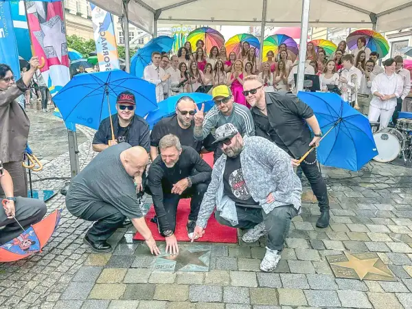 Grupa mężczyzn, prawdopodobnie zespół, odsłania gwiazdę z napisem "PIERSI BAŁKANICA" na chodniku. W tle kolorowe parasole i publiczność.