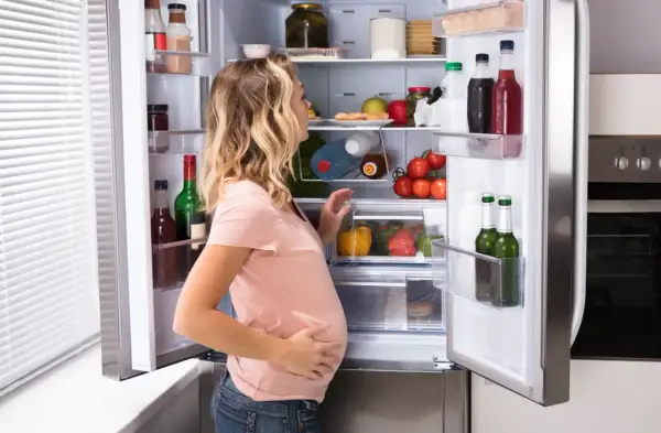 Ciężarna kobieta przegląda lodówkę, zastanawiając się, czy w ciąży można jeść ostre. W środku warzywa, owoce i napoje.