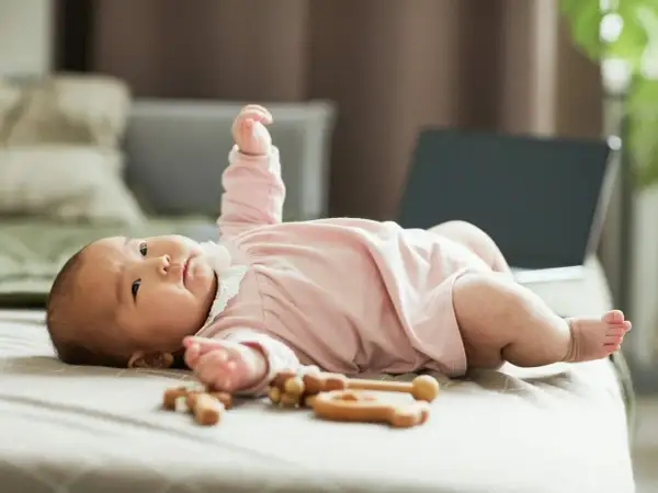 Urocze dziecko patrzy w jeden punkt i się śmieje, leżąc na łóżku obok drewnianych zabawek.