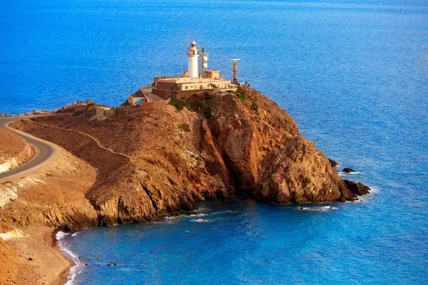 Faro en un acantilado rocoso con el mar azul de fondo. Un camino serpentea hacia la cima. ¡Un lugar perfecto para ver Cabo de Gata!