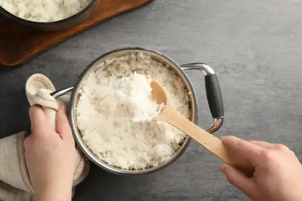 Hände halten einen Topf mit frisch gekochtem Reis. Eine hölzerne Kelle hebt eine Portion Reis heraus, perfekt auf welcher Stufe Reis kochen.