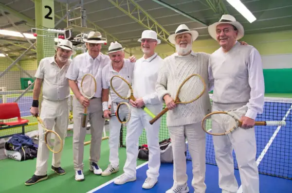 Sex äldre herrar i hattar och vita kläder poserar med tennisracketar, redo för en match i stil med Gustaf V:s tennis.