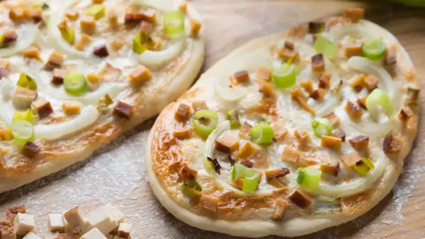 Zwei knusprige, vegetarische Flammkuchen mit Zwiebeln, Frühlingszwiebeln und würzigen Tofuwürfeln, bestreut mit etwas Mehl.