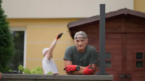 Mężczyzna w czapce i rękawiczkach opiera się o mur. W tle inny mężczyzna maluje drewnianą szopę. Jak zgłosić się do polowania na ogród?