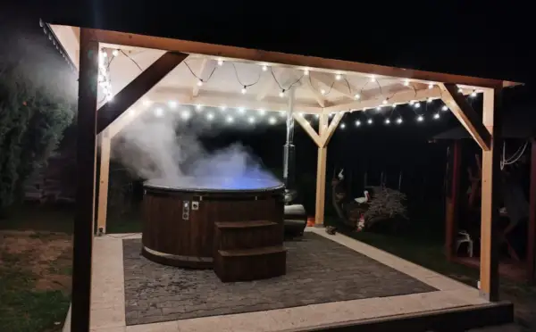 Drewniana beczka z gorącą wodą, z której unosi się para, pod zadaszeniem ozdobionym lampkami. Idealne miejsce na relaks w stylu sauna fińska.