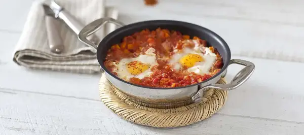 Un plat d'oeuf à la tomate mijoté dans une poêle, servi sur un dessous de plat en osier.