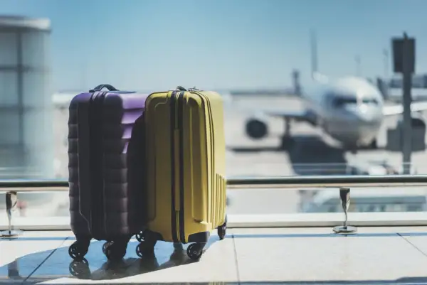 Zwei Koffer, lila und gelb, stehen am Flughafen vor einem Flugzeug.