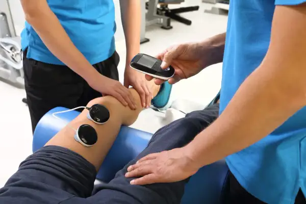 Fizjoterapeuta wykonuje zabieg fonoforezy na kolanie pacjenta, używając aparatu i elektrod.