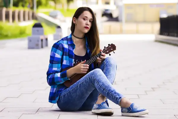 Dziewczyna w kratę i jeansach pokazuje, jak trzymać ukulele, siedząc na chodniku.