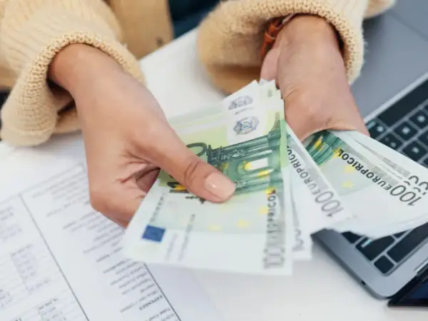 Mani che contano banconote da 100 euro, forse per la divisione conto corrente eredi. Documenti e laptop sullo sfondo.