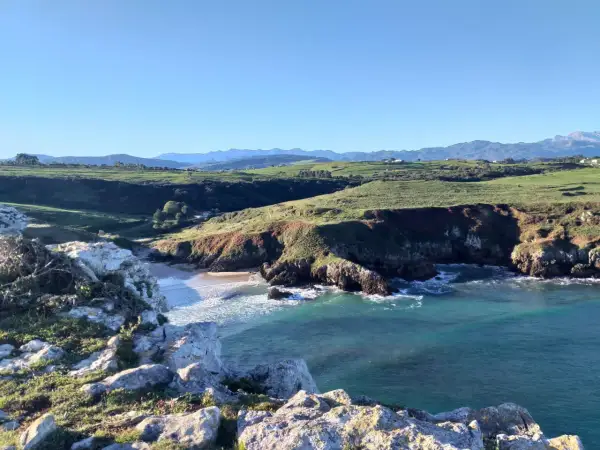 Acantilados y mar turquesa, ideales para rutas senderismo San Vicente de la Barquera. Paisaje costero con verdes colinas y montañas al fondo.
