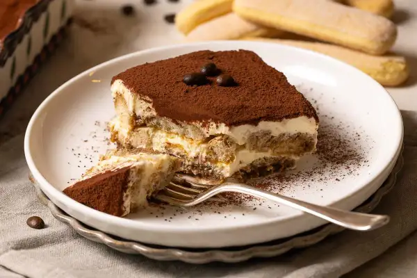 Ein Stück Tiramisu, zubereitet nach einem einfachen tiramisu rezept, liegt auf einem Teller. Kakaopulver bestäubt die cremige Schicht und die Löffelbiskuits.