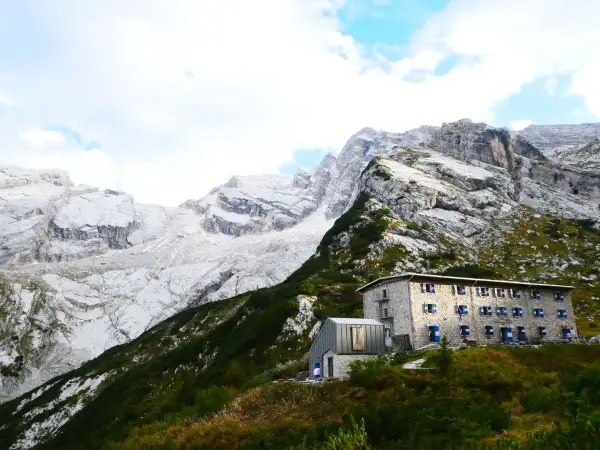 Un rifugio in pietra con persiane blu si staglia su un pendio verdeggiante, sotto imponenti montagne rocciose. Un esempio di rifugi in Veneto.