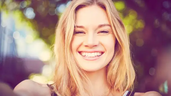 Strahlendes Lächeln einer jungen Frau mit blonden Haaren, die eine natürliche Frisur trägt.
