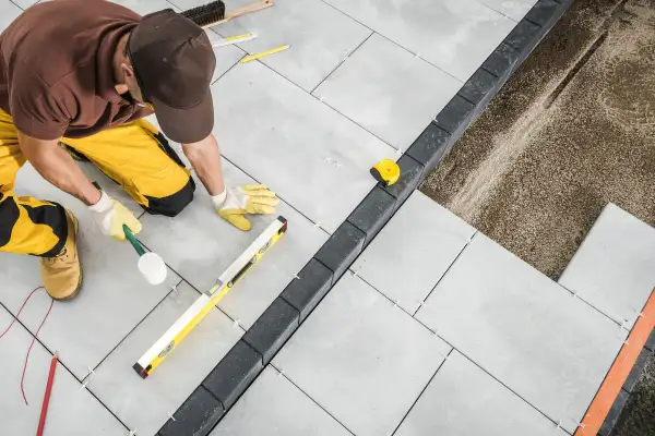 Operaio con martello di gomma e livella esegue la posa a secco di pavimenti esterni in lastre grigie, creando un bordo con pietre scure.