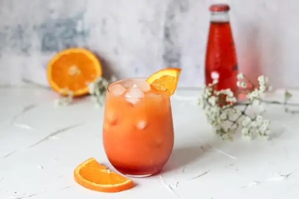 Ein Glas Aperitif mit Eis und Orangenscheibe, daneben eine halbe Orange und eine rote Flasche.