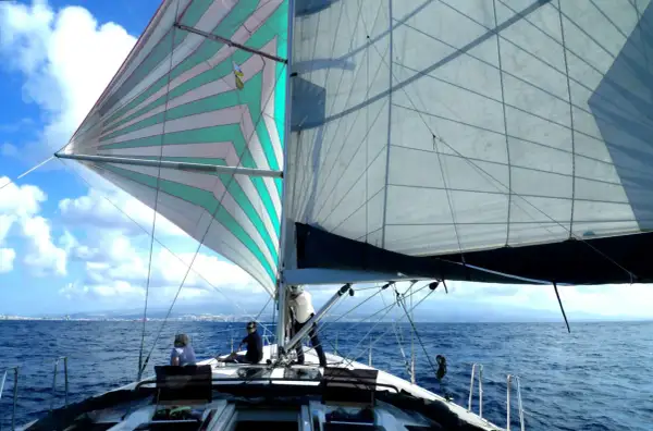 Vela colorata e bianca gonfie al vento, con persone a bordo pronte a mettersi alla cappa. Mare blu e cielo con nuvole bianche.
