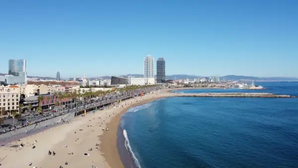 Słoneczna plaża Barceloneta z turkusową wodą, palmami i nowoczesnymi budynkami w tle.