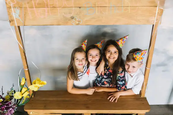 Dzieci w czapkach urodzinowych pozują przy drewnianej konstrukcji z napisem "Happy Birthday". To świetny pomysł, jak zrobić fotobudkę z kartonu na przyjęcie!