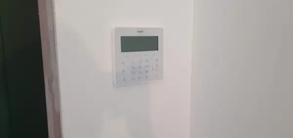 Biały panel alarmowy z klawiaturą numeryczną i ekranem na białej ścianie.