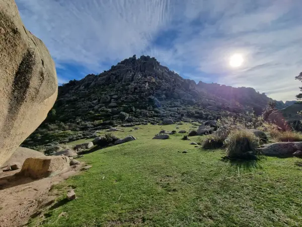 Majestuosa montaña rocosa en la ruta Soto del Real, con un prado verde y el sol brillando en un cielo azul con nubes.