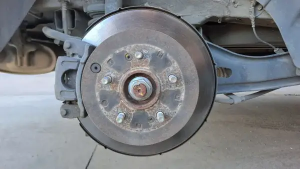 Gros plan sur un disque de frein usé, rouillé et marqué par l'usage. L'étrier de frein est visible, ainsi que des éléments de suspension.