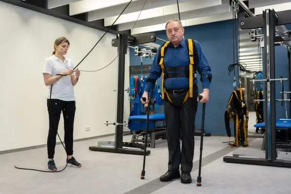 Starszy mężczyzna w specjalistycznej uprzęży i z kulami ćwiczy pod okiem fizjoterapeutki. To kluczowy etap rehabilitacji neurologicznej.