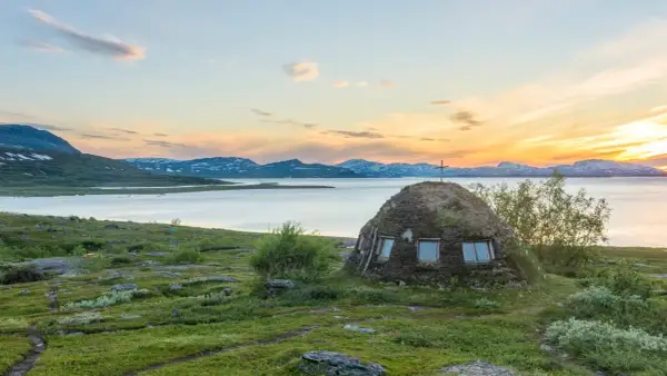En grästorvstuga med kors på toppen vid en sjö i Padjelanta nationalpark, Sverige, under en solnedgång.