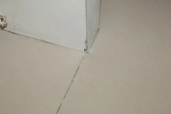 Uszkodzona dylatacja posadzki w rogu pomieszczenia, widoczne pęknięcia i ubytki materiału.