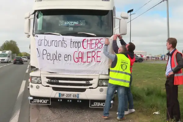 Des routiers accrochent une banderole sur un camion : "Carburants trop CHERS, Peuple en GALERE". Ils expriment leur mécontentement face au prix du carburant.