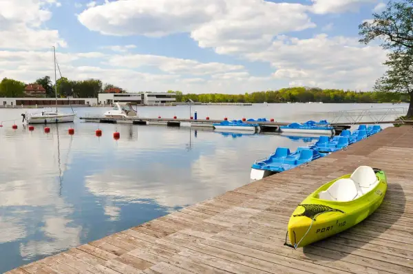 Iława atrakcje: pomost z kajakiem i rzędem rowerów wodnych na spokojnym jeziorze, w tle żaglówka i budynki portowe.