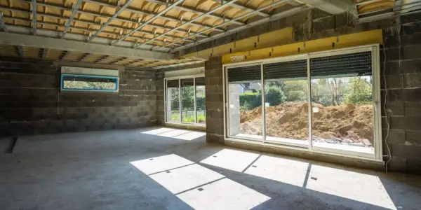 Wnętrze w budowie z betonową podłogą i ścianami. Widoczna konstrukcja sufitu i okna z widokiem na plac budowy. Powstaje nowa ściana działowa.