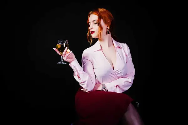 Donna con capelli rossi tiene un calice di vino da meditazione, con un'espressione pensierosa. Indossa una camicetta rosa e una gonna bordeaux.