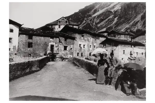 Un villaggio di montagna immortalato da Giovanni Verga fotografo. Coppie eleganti passeggiano con ombrelli, sullo sfondo case in pietra e pendii scoscesi.