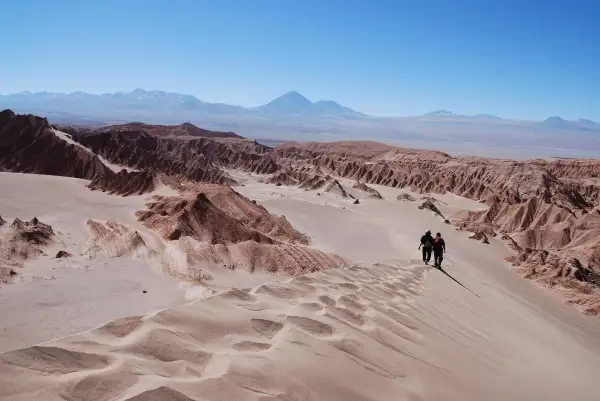 Dos excursionistas caminan por una duna en el desierto de Atacama, con formaciones rocosas y montañas al fondo bajo un cielo azul.