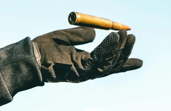 Una mano con guantes tácticos sostiene un cartucho dorado contra un cielo azul claro.