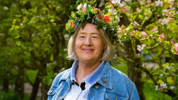 Glad kvinna med blomsterkrans, Bettan byvald, ler i solsken bland blommande träd.