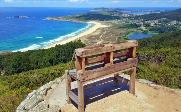 Banco de madera en un monte ventoso con vistas a una playa, el mar azul y un lago.