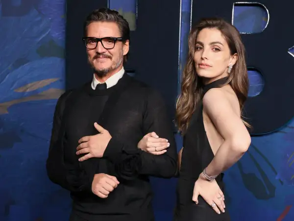 Pedro Pascal y una mujer elegante posan juntos. El actor, con gafas y un suéter negro, luce un cabestrillo. Ella, con un vestido negro y joyas brillantes, sonríe.