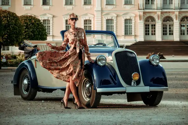 Elegancka kobieta w kwiecistej sukience retro z lat 20. pozuje przy zabytkowym kabriolecie przed pałacem.