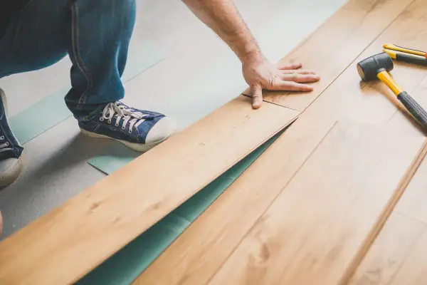 Pose d'un parquet flottant, une main ajuste une lame. Outil et maillet à proximité.