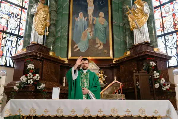 Ksiądz w zielonej szacie błogosławi wiernych. W tle obraz Jezusa na krzyżu. To moment święcenia wody w kościele.
