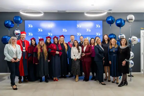 Grupa absolwentów i wykładowców świętuje 30-lecie swojej alma mater. Wszyscy ubrani są w togi i birety, a w tle wiszą balony.