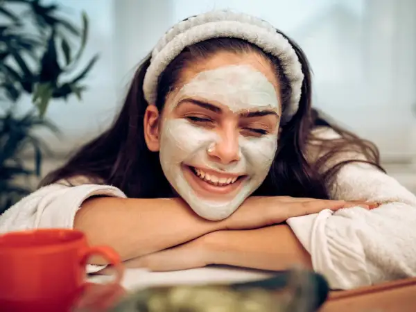 Lächelnde Frau mit Gesichtsmaske, die sich fragt: nach Gesichtsmaske eincremen oder nicht? Entspannter Moment mit rotem Becher.