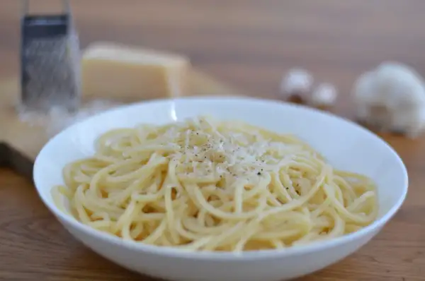 Ein Teller mit dampfenden Spaghetti, bestreut mit frisch geriebenem Parmesan. Dazu ein Stück Parmesan, eine Reibe und Knoblauch – perfekt für ein authentisches italienisches Gericht.