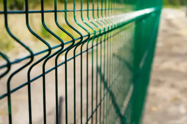 Zielone ogrodzenie z siatki bez podmurówki, z charakterystycznymi przetłoczeniami, biegnie wzdłuż piaszczystej ścieżki.
