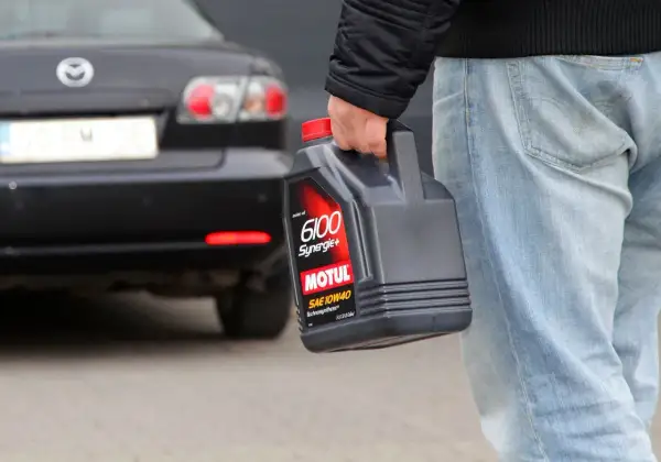 Mężczyzna niesie kanister z olejem silnikowym Motul 6100 Synergie+ SAE 10W40. Za nim stoi czarny samochód.