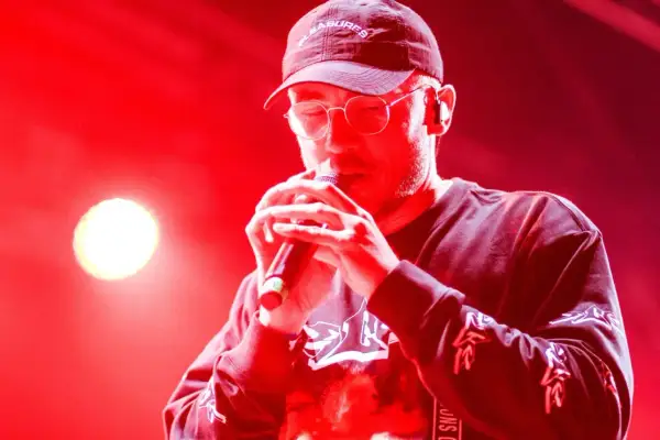 Raper w czapce i okularach śpiewa do mikrofonu. Jego album zdobył Fryderyka w kategorii album roku (hip-hop).