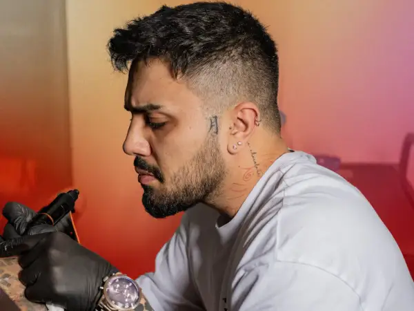 Un homme se fait tatouer le cou, un tatouage homme cou discret avec des motifs rouges. Il porte des gants noirs et une montre.